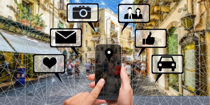 Graphic of a city street with people congregating. In the foreground, a hand operating a cell phone with geometric lines covering the screen and bubbles with icons for camera, email, social media, and rideshare. Represents digital communication, 5g networking