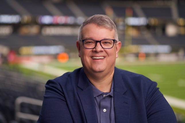 A man with glasses and short cropped gray hair smiles from a seat in a baseball stadium.